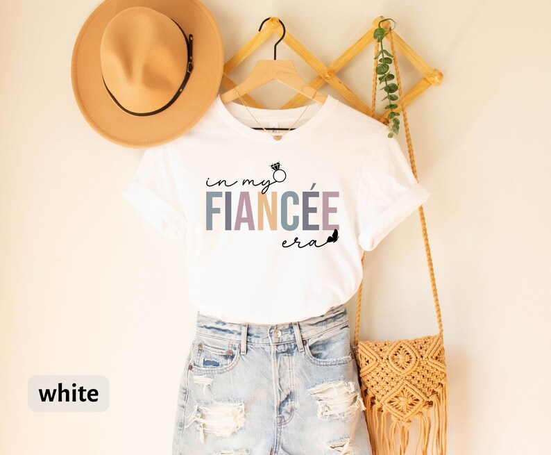 May include: White t-shirt with the text "in my fiancee era" in colorful letters with a ring graphic above the word "fiancee".