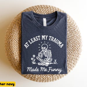 K&ouml;nnte beinhalten: Heather-Navy-T-Shirt mit dem Text "AT LEAST MY TRAUMA Made Me Funny" und einer wei&szlig;en Skelettgrafik. Das Skelett sitzt und h&auml;lt eine Tasse, mit Blumen. Das Shirt liegt auf einer gewebten, kreisf&ouml;rmigen Oberfl&auml;che.