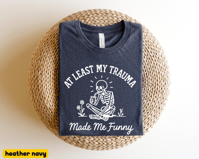 K&ouml;nnte beinhalten: Heather-Navy-T-Shirt mit dem Text "AT LEAST MY TRAUMA Made Me Funny" und einer wei&szlig;en Skelettgrafik. Das Skelett sitzt und h&auml;lt eine Tasse, mit Blumen. Das Shirt liegt auf einer gewebten, kreisf&ouml;rmigen Oberfl&auml;che.