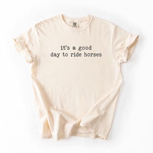 May include: A beige t-shirt with the text "it's a good day to ride horses" printed in black.