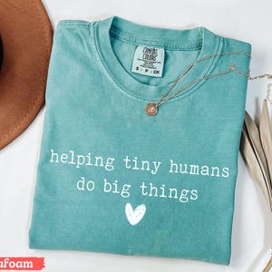 May include: Seafoam green t-shirt with the text "helping tiny humans do big things" and a white heart graphic. A gold necklace with a pendant is draped across the shirt. The shirt is folded on a white surface.