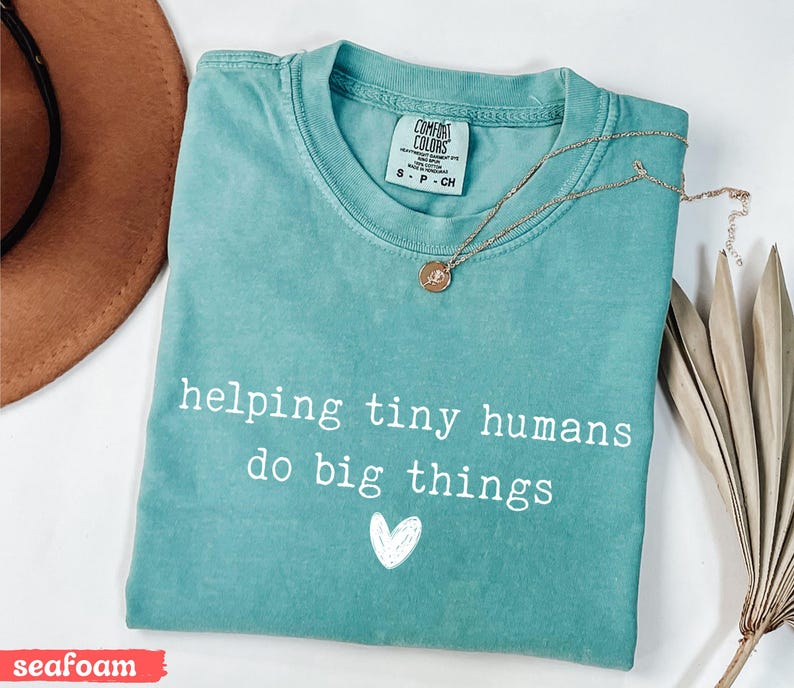 May include: Seafoam green t-shirt with the text "helping tiny humans do big things" and a white heart graphic. A gold necklace with a pendant is draped across the shirt. The shirt is folded on a white surface.
