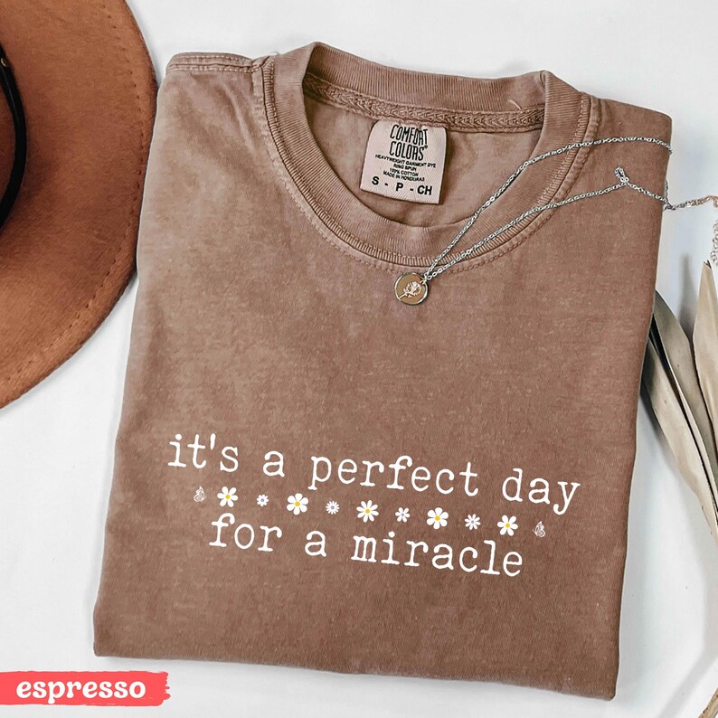 May include: A brown t-shirt with the text "it's a perfect day for a miracle" and small white flower graphics. The shirt is folded and laid flat on a white surface.