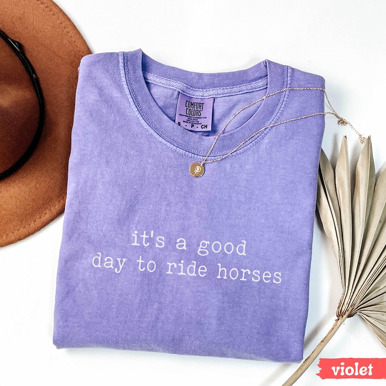May include: A purple t-shirt with the text "it's a good day to ride horses" in white. The shirt is folded and laid on a white surface.