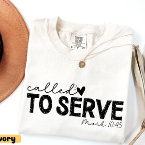 Pu&ograve; includere: T-shirt color avorio con le parole "called TO SERVE" in nero, insieme a un simbolo a forma di cuore. Il testo "Mark 10:45" &egrave; in basso. Una collana d'oro e un cappello marrone sono visibili sullo sfondo. La maglietta &egrave; etichettata "Comfort Colors".