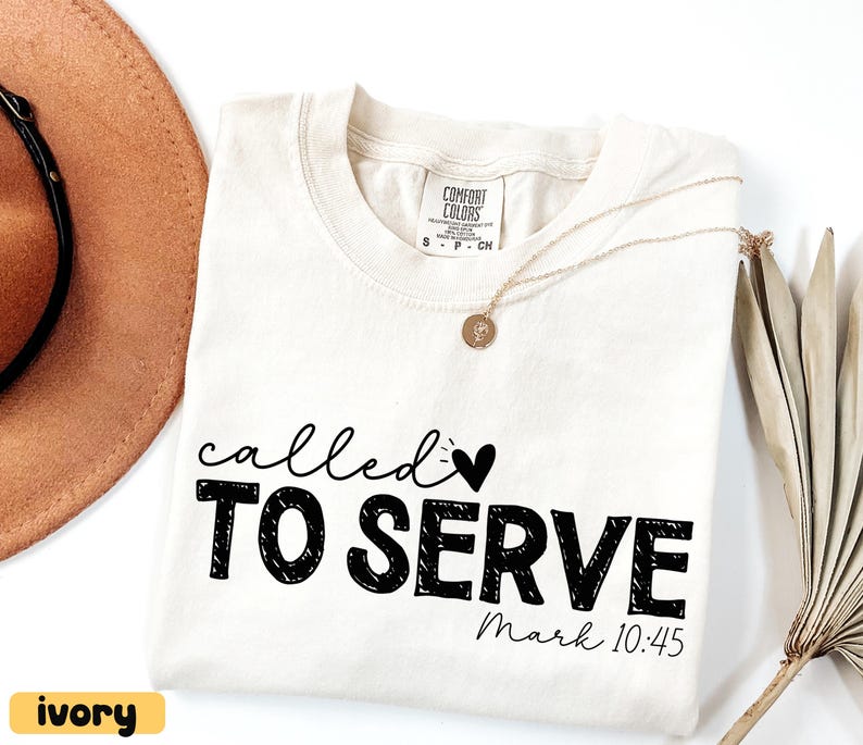 Pu&ograve; includere: T-shirt color avorio con le parole "called TO SERVE" in nero, insieme a un simbolo a forma di cuore. Il testo "Mark 10:45" &egrave; in basso. Una collana d'oro e un cappello marrone sono visibili sullo sfondo. La maglietta &egrave; etichettata "Comfort Colors".