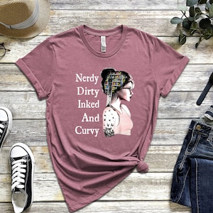 Op de afbeelding: Een mauve t-shirt met een afbeelding van een vrouw met boeken op haar hoofd en een tatoeage op haar arm. De tekst op het shirt luidt "Nerdy Dirty Inked And Curvy".