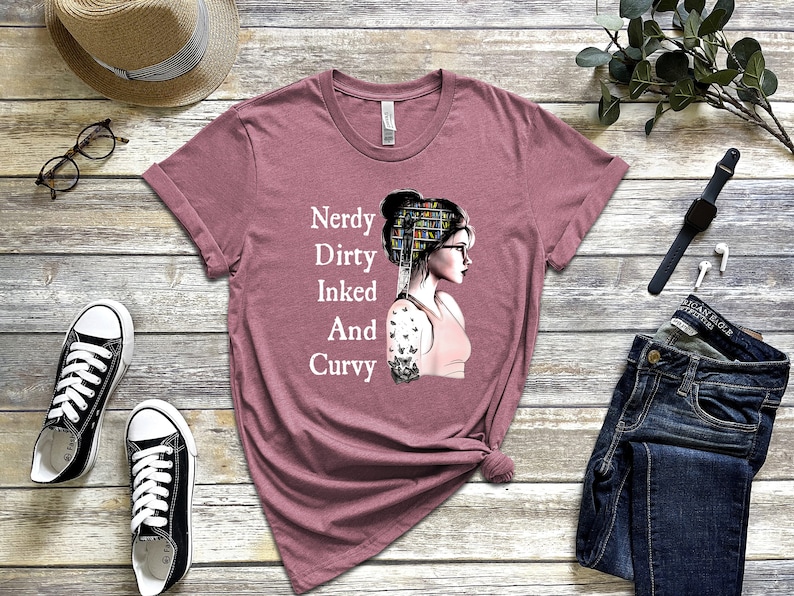 Op de afbeelding: Een mauve t-shirt met een afbeelding van een vrouw met boeken op haar hoofd en een tatoeage op haar arm. De tekst op het shirt luidt "Nerdy Dirty Inked And Curvy".