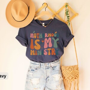 May include: A navy blue t-shirt with the text "Motherhood is my ministry" in colorful, retro-style lettering. The shirt is displayed on a hanger with a straw hat and a woven bag.