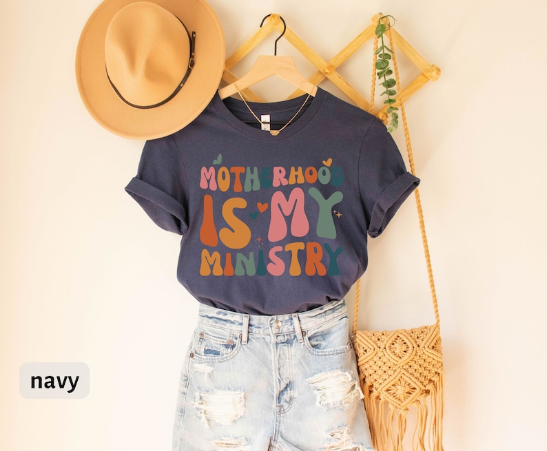 May include: A navy blue t-shirt with the text "Motherhood is my ministry" in colorful, retro-style lettering. The shirt is displayed on a hanger with a straw hat and a woven bag.