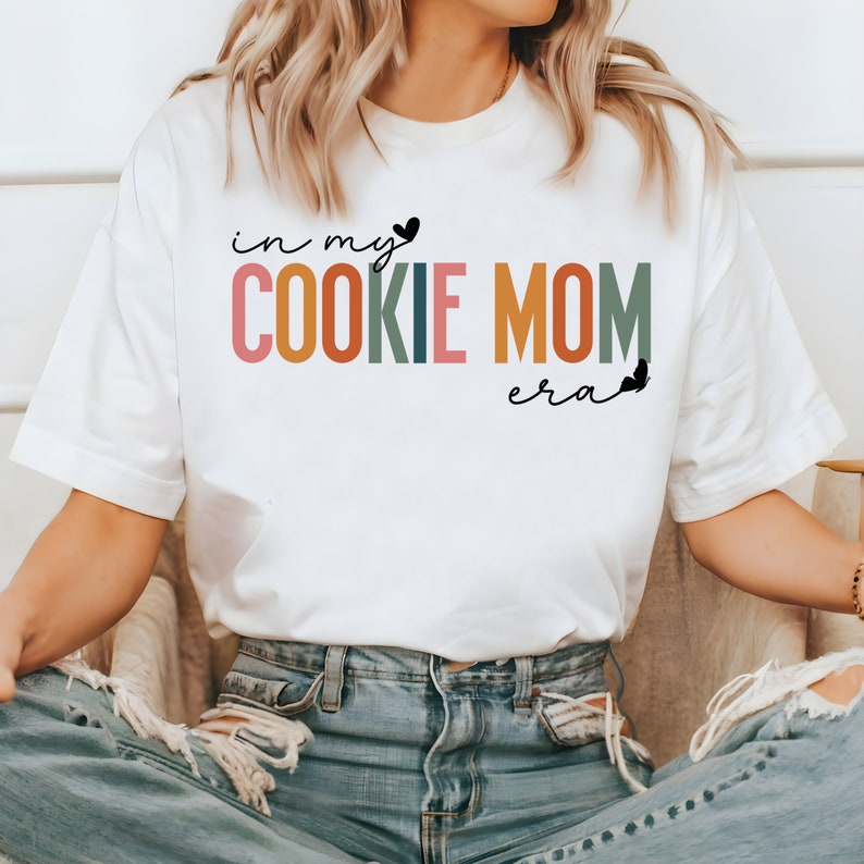 May include: White t-shirt with the text "in my cookie mom era" in rainbow colors with a small heart and butterfly graphic.