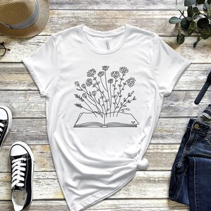 K&ouml;nnte beinhalten: Wei&szlig;es T-Shirt mit einer schwarzen Linienzeichnung eines offenen Buches, aus dem Blumen wachsen.