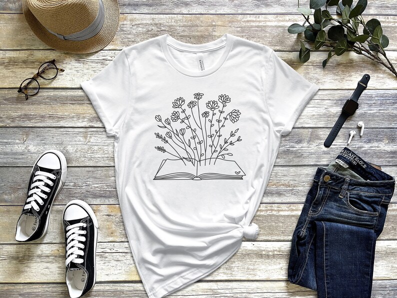 K&ouml;nnte beinhalten: Wei&szlig;es T-Shirt mit einer schwarzen Linienzeichnung eines offenen Buches, aus dem Blumen wachsen.