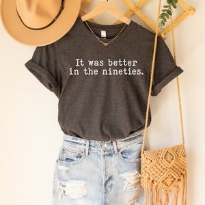 K&ouml;nnte beinhalten: Ein dunkelgraues T-Shirt mit dem Text "It was better in the nineties." in wei&szlig; gedruckt. Das Shirt h&auml;ngt an einem Holzb&uuml;gel mit einem Strohhut und einer gewebten Tasche.
