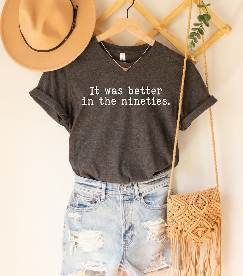 K&ouml;nnte beinhalten: Ein dunkelgraues T-Shirt mit dem Text "It was better in the nineties." in wei&szlig; gedruckt. Das Shirt h&auml;ngt an einem Holzb&uuml;gel mit einem Strohhut und einer gewebten Tasche.