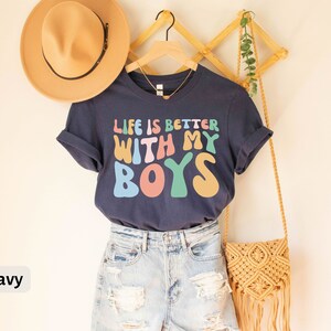 K&ouml;nnte beinhalten: Ein marineblaues T-Shirt mit dem Text "Life is better with my boys" in bunten, Retro-Lettern. Das Shirt ist an einem Kleiderb&uuml;gel mit einem Strohhut und einer geflochtenen Tasche aufgeh&auml;ngt.