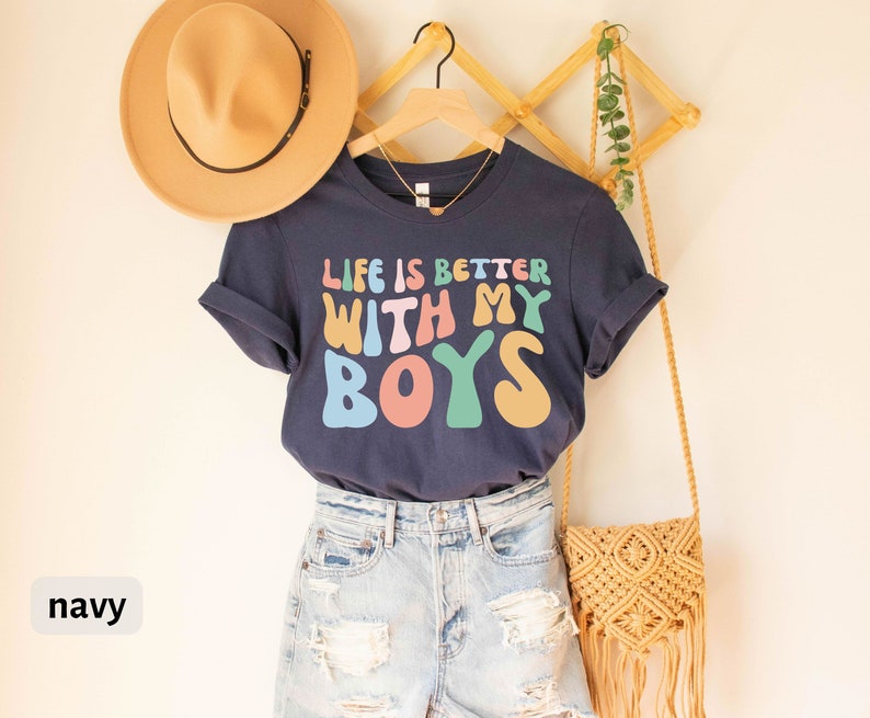 K&ouml;nnte beinhalten: Ein marineblaues T-Shirt mit dem Text "Life is better with my boys" in bunten, Retro-Lettern. Das Shirt ist an einem Kleiderb&uuml;gel mit einem Strohhut und einer geflochtenen Tasche aufgeh&auml;ngt.