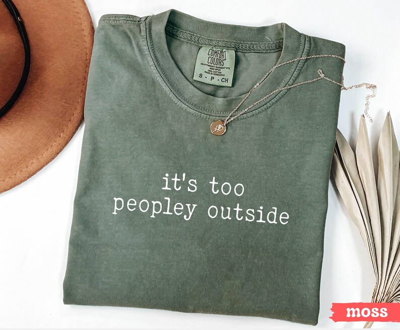 May include: A green t-shirt with the text "it's too peopley outside" printed in white. The shirt is folded and laid flat on a white surface.
