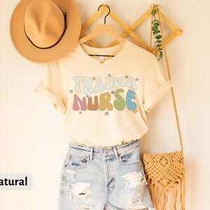 May include: A beige t-shirt with the text "Trauma Nurse" in rainbow colors and stars. The shirt is hanging on a wooden hanger with a straw bag and a hat.