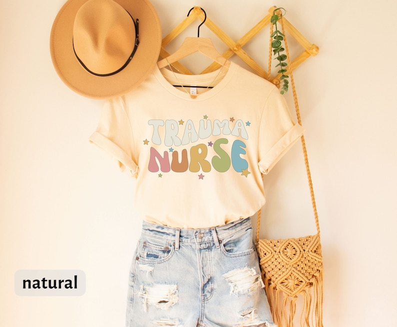 May include: A beige t-shirt with the text "Trauma Nurse" in rainbow colors and stars. The shirt is hanging on a wooden hanger with a straw bag and a hat.
