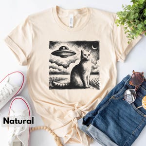 May include: A beige t-shirt with a black and white illustration of a cat looking up at a flying saucer. The illustration is in a vintage style.