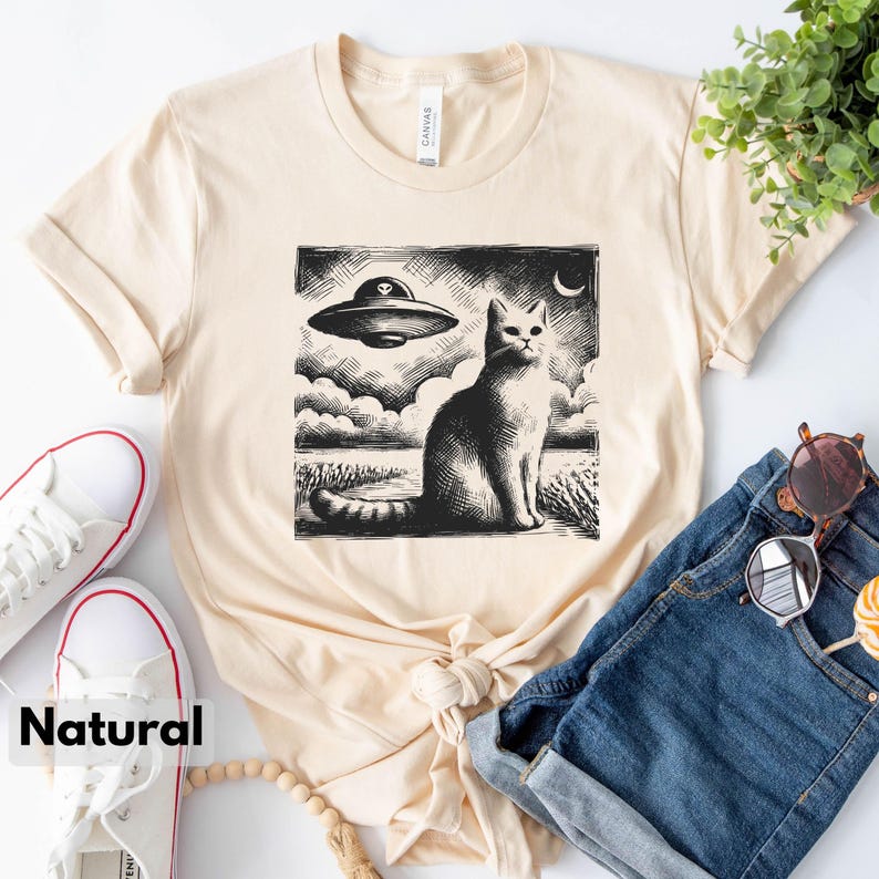 May include: A beige t-shirt with a black and white illustration of a cat looking up at a flying saucer. The illustration is in a vintage style.