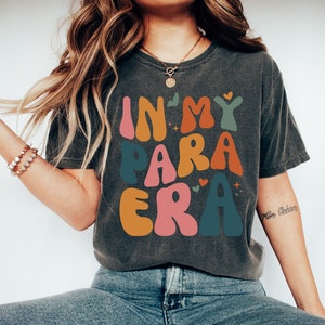 May include: A dark gray t-shirt with the text "In My Para Era" in colorful, retro-style lettering. The letters are outlined in white and have a slightly distressed look.