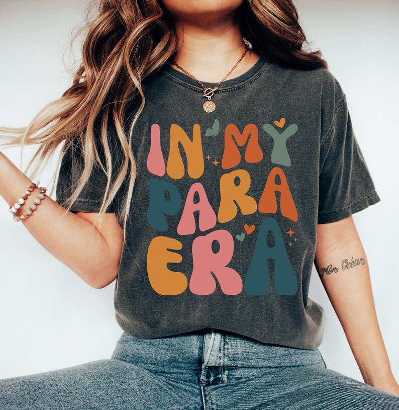 May include: A dark gray t-shirt with the text "In My Para Era" in colorful, retro-style lettering. The letters are outlined in white and have a slightly distressed look.