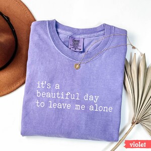 May include: A purple t-shirt with the text "it's a beautiful day to leave me alone" in white. The shirt is folded and laid flat on a white surface.