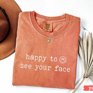 May include: A coral colored t-shirt with the text "happy to see your face" and a smiley face in white. The shirt is made of a soft, comfortable material.