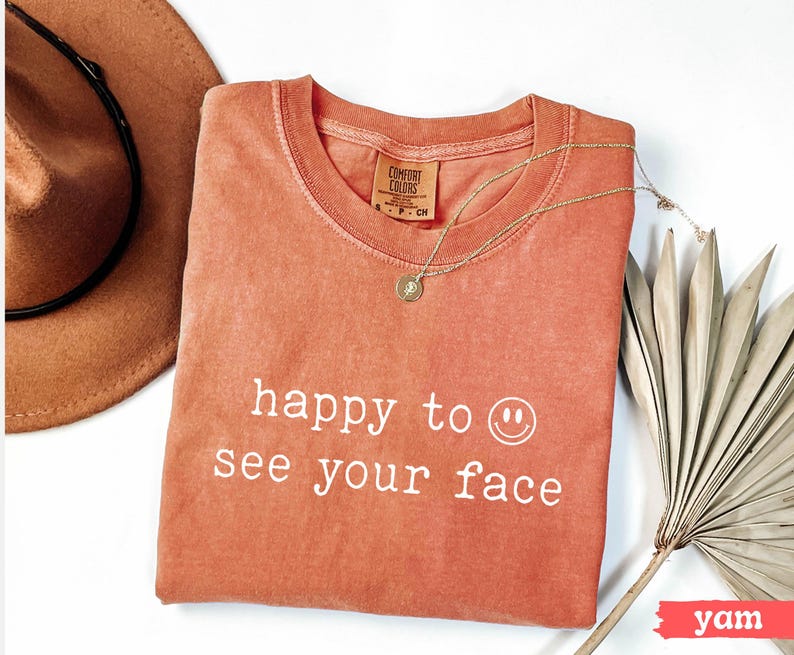May include: A coral colored t-shirt with the text "happy to see your face" and a smiley face in white. The shirt is made of a soft, comfortable material.