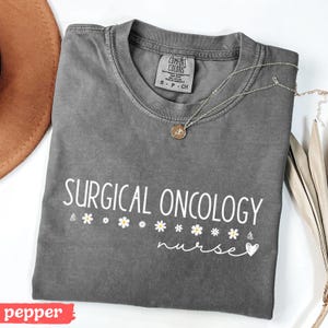 May include: A gray t-shirt with the text "SURGICAL ONCOLOGY nurse" in white lettering. The shirt has a small heart and daisy accents.