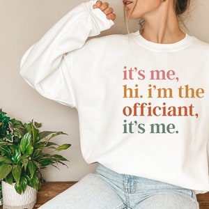 May include: White sweatshirt with the text "it's me, hi. i'm the officiant, it's me." printed in pink, orange, and green.
