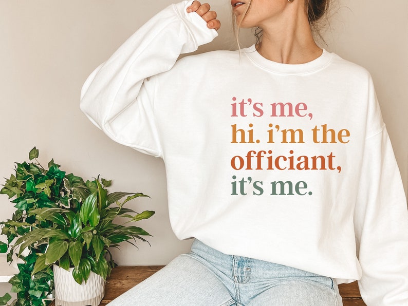 May include: White sweatshirt with the text "it's me, hi. i'm the officiant, it's me." printed in pink, orange, and green.
