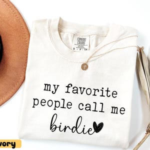 May include: Ivory Comfort Colors t-shirt with the text "my favourite people call me birdie" in black. A gold necklace with a pendant and a brown hat are visible. A dried palm leaf is on the right.