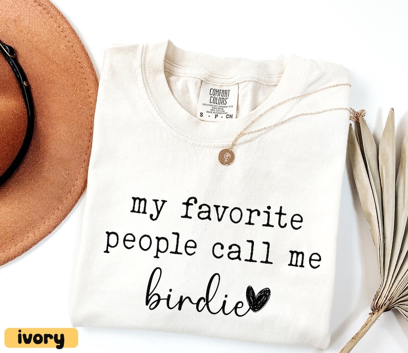 May include: Ivory Comfort Colors t-shirt with the text "my favourite people call me birdie" in black. A gold necklace with a pendant and a brown hat are visible. A dried palm leaf is on the right.