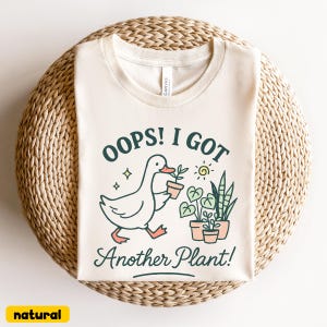 May include: A cream-colored t-shirt with the text "OOPS! I GOT Another Plant!" and a graphic of a duck carrying a potted plant. The shirt also features illustrations of plants and a sun.