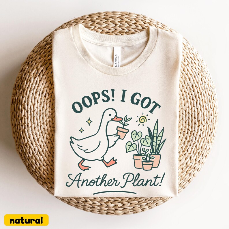 May include: A cream-colored t-shirt with the text "OOPS! I GOT Another Plant!" and a graphic of a duck carrying a potted plant. The shirt also features illustrations of plants and a sun.