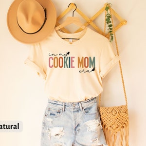 May include: A natural colored t-shirt with the text "in my cookie mom era" in rainbow colors. The shirt is hanging on a wooden hanger with a straw bag and a hat.