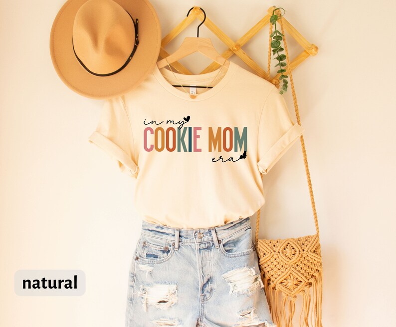 May include: A natural colored t-shirt with the text "in my cookie mom era" in rainbow colors. The shirt is hanging on a wooden hanger with a straw bag and a hat.