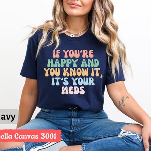 May include: A navy blue t-shirt with the text "If you're happy and you know it, it's your meds" in rainbow colors. The shirt is made by Bella Canvas and is a size 3001.