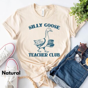 May include: A beige t-shirt with a blue graphic of a goose carrying books. The text "Silly Goose Teacher Club" is printed on the shirt.
