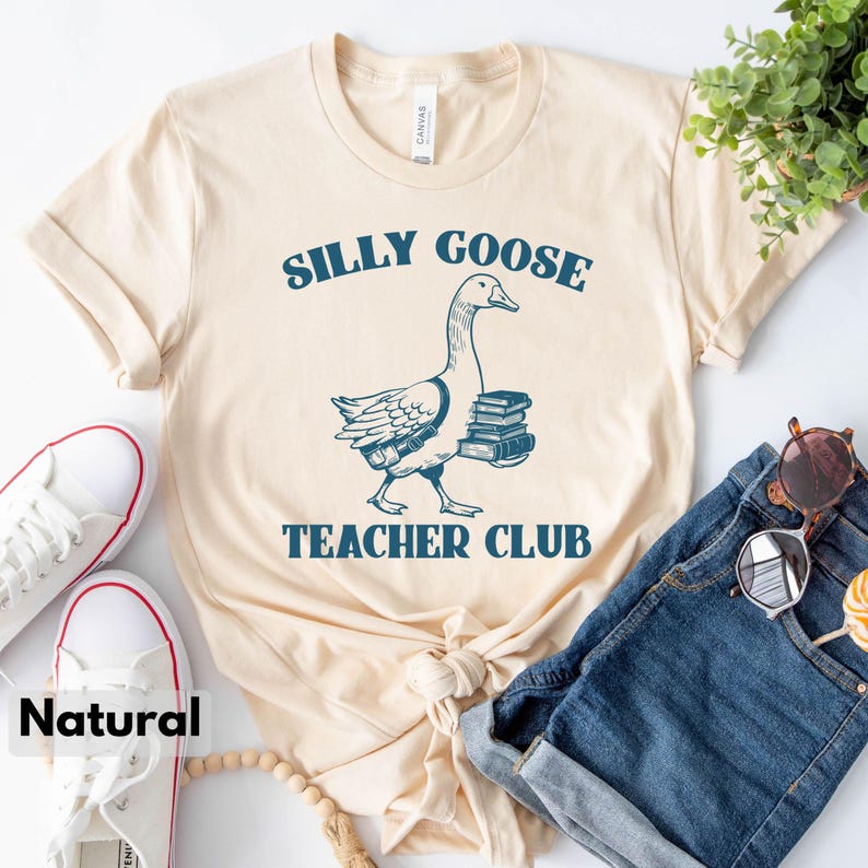 May include: A beige t-shirt with a blue graphic of a goose carrying books. The text "Silly Goose Teacher Club" is printed on the shirt.