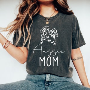 K&ouml;nnte beinhalten: Ein dunkelgraues T-Shirt mit einer wei&szlig;en Umrandung eines Australian Shepherd Hundes und dem Text "Aussie MOM" auf der Vorderseite.