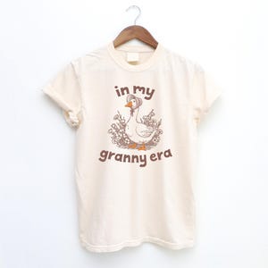 May include: Cream-colored t-shirt with a graphic of a goose wearing a bonnet, surrounded by flowers. The text "in my granny era" is printed in brown above and below the goose illustration. The shirt is hanging on a wooden hanger.