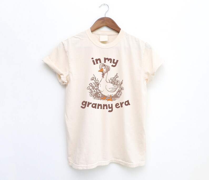 May include: Cream-colored t-shirt with a graphic of a goose wearing a bonnet, surrounded by flowers. The text "in my granny era" is printed in brown above and below the goose illustration. The shirt is hanging on a wooden hanger.