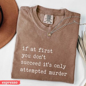 Op de afbeelding: Espresso-kleurig Comfort Colors T-shirt met de witte quote "if at first you don't succeed it's only attempted murder". Een zilveren ketting met hanger ligt op het shirt. Maat S.