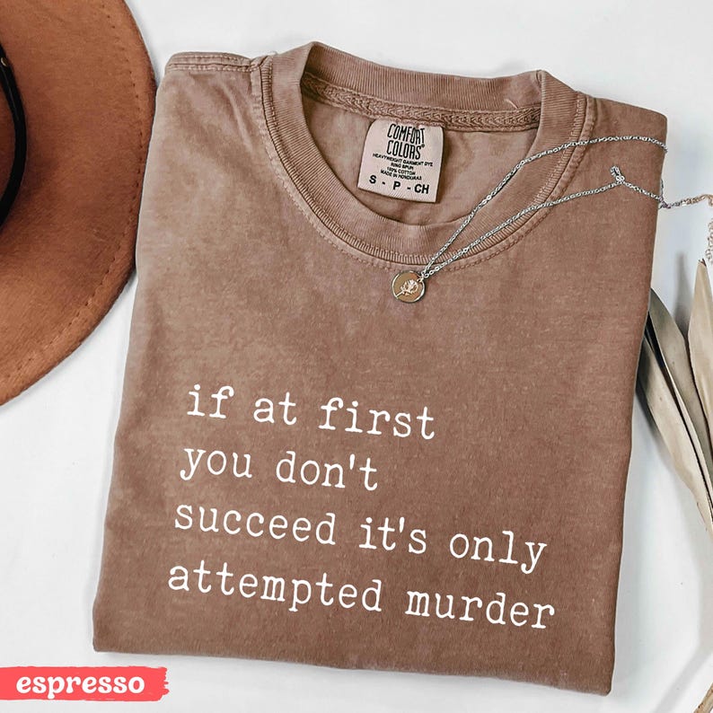 Op de afbeelding: Espresso-kleurig Comfort Colors T-shirt met de witte quote "if at first you don't succeed it's only attempted murder". Een zilveren ketting met hanger ligt op het shirt. Maat S.