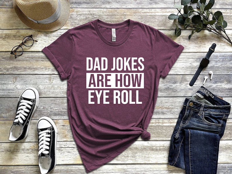 May include: A maroon t-shirt with white text that reads "Dad Jokes Are How Eye Roll".