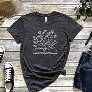 K&ouml;nnte beinhalten: Ein dunkelgraues T-Shirt mit einer wei&szlig;en Umrandung eines Buches, aus dem Blumen wachsen.